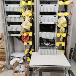 FiberOptic - Dwa panele krosowe w szafie rack, z żółtymi elementami prowadzenia kabli, kable światłowodowe w folii, skrzynka z akcesoriami i dokumentacja na podłodze, stół roboczy z dodatkowym panelem krosowym.