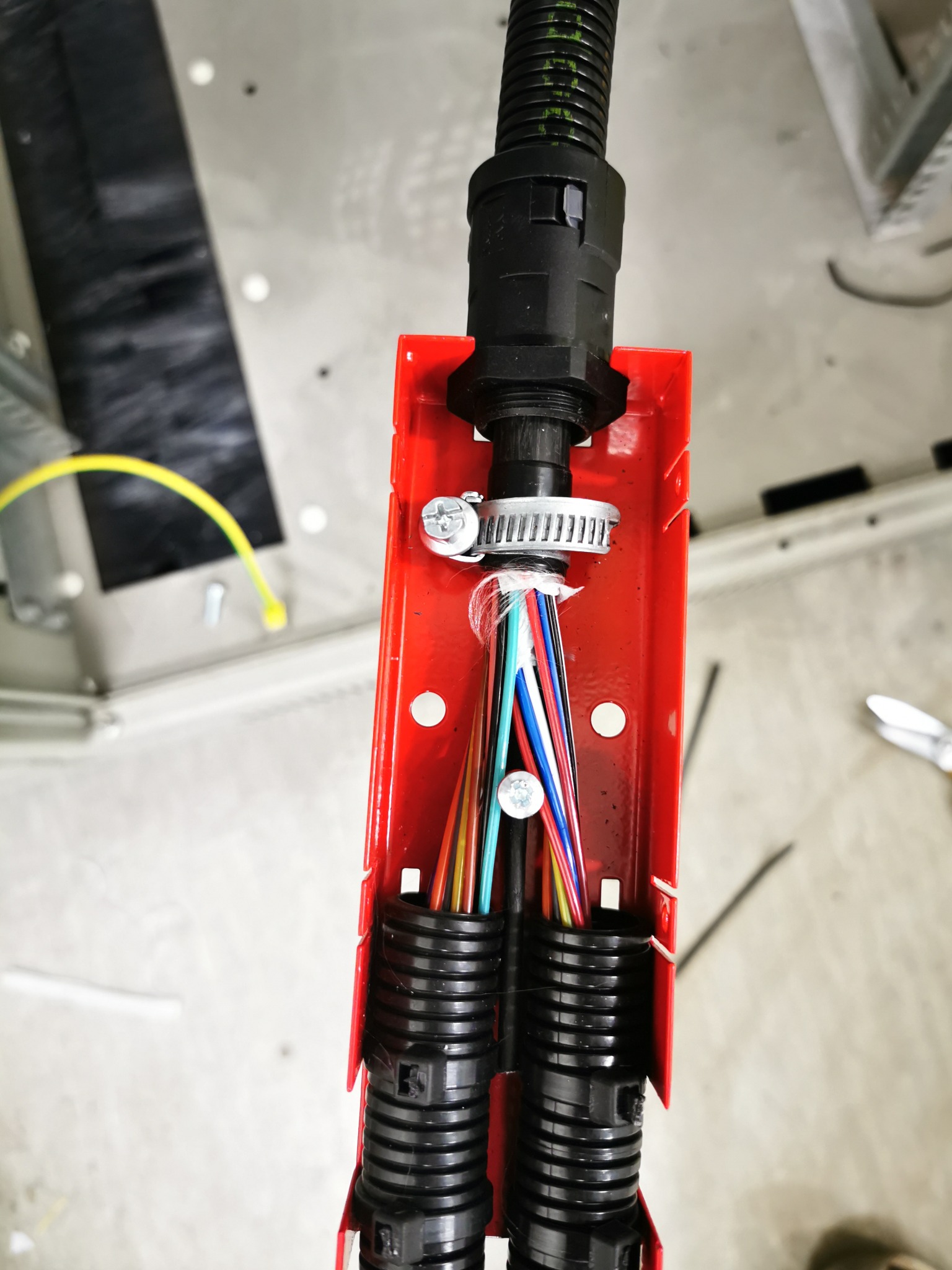 Widok z góry na kable światłowodowe w czarnej karbowanej rurce, przechodzące przez czerwoną skrzynkę montażową zabezpieczone opaską zaciskową, z widocznymi kolorowymi włóknami.