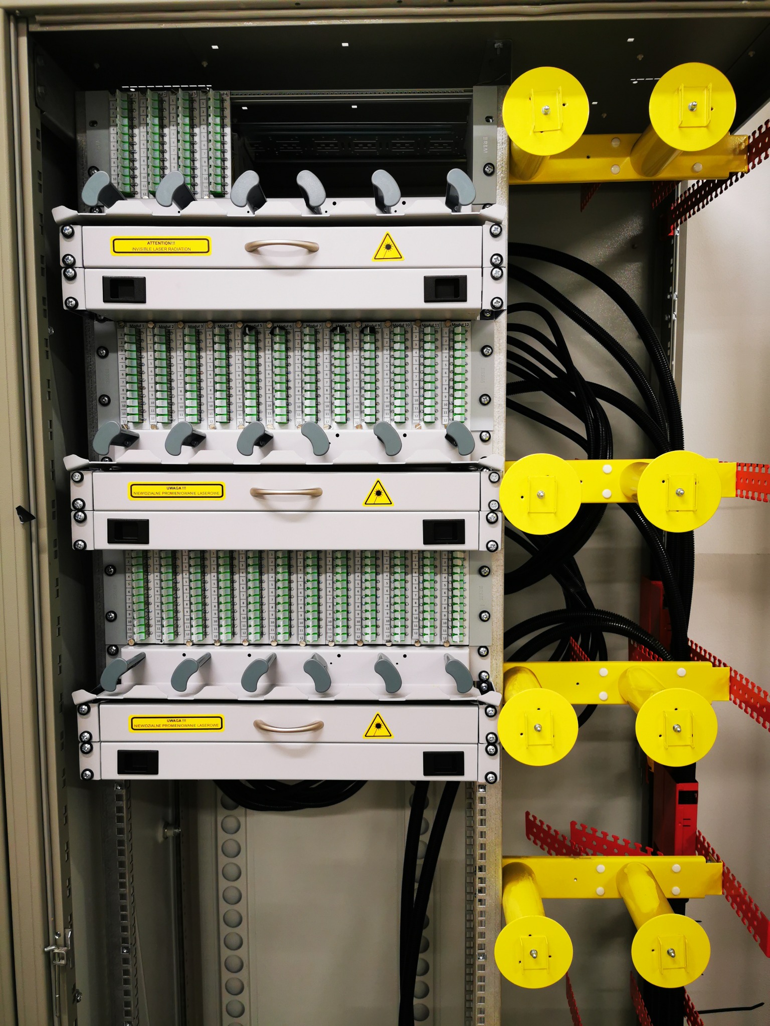 Panel krosowniczy światłowodowy w szafie rack z żółtymi uchwytami kablowymi, widoczne moduły z zielonymi wkładkami i oznaczeniami, ostrzeżenia o promieniowaniu laserowym.