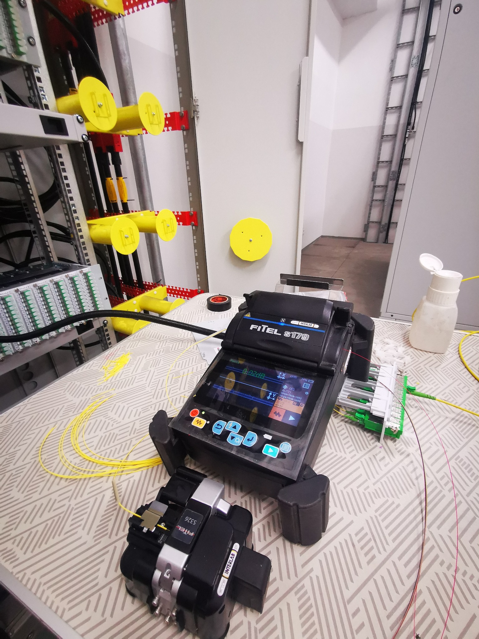 Sprzęt Fitel S178 do spawania światłowodów na stole z widocznym panelem krosowniczym, żółtymi prowadnicami kablowymi i pojemnikiem z płynem do czyszczenia światłowodów.