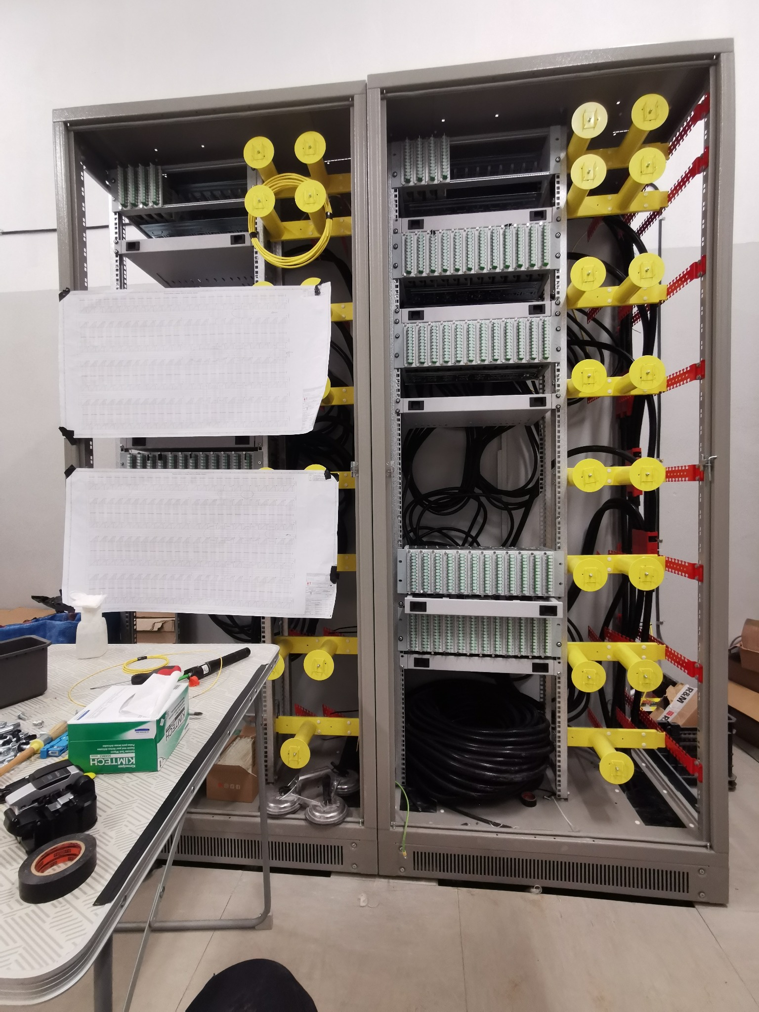 Dwie szafy telekomunikacyjne z panelami krosowymi i uporządkowanymi kablami, na stole obok narzędzia i schematy instalacji.