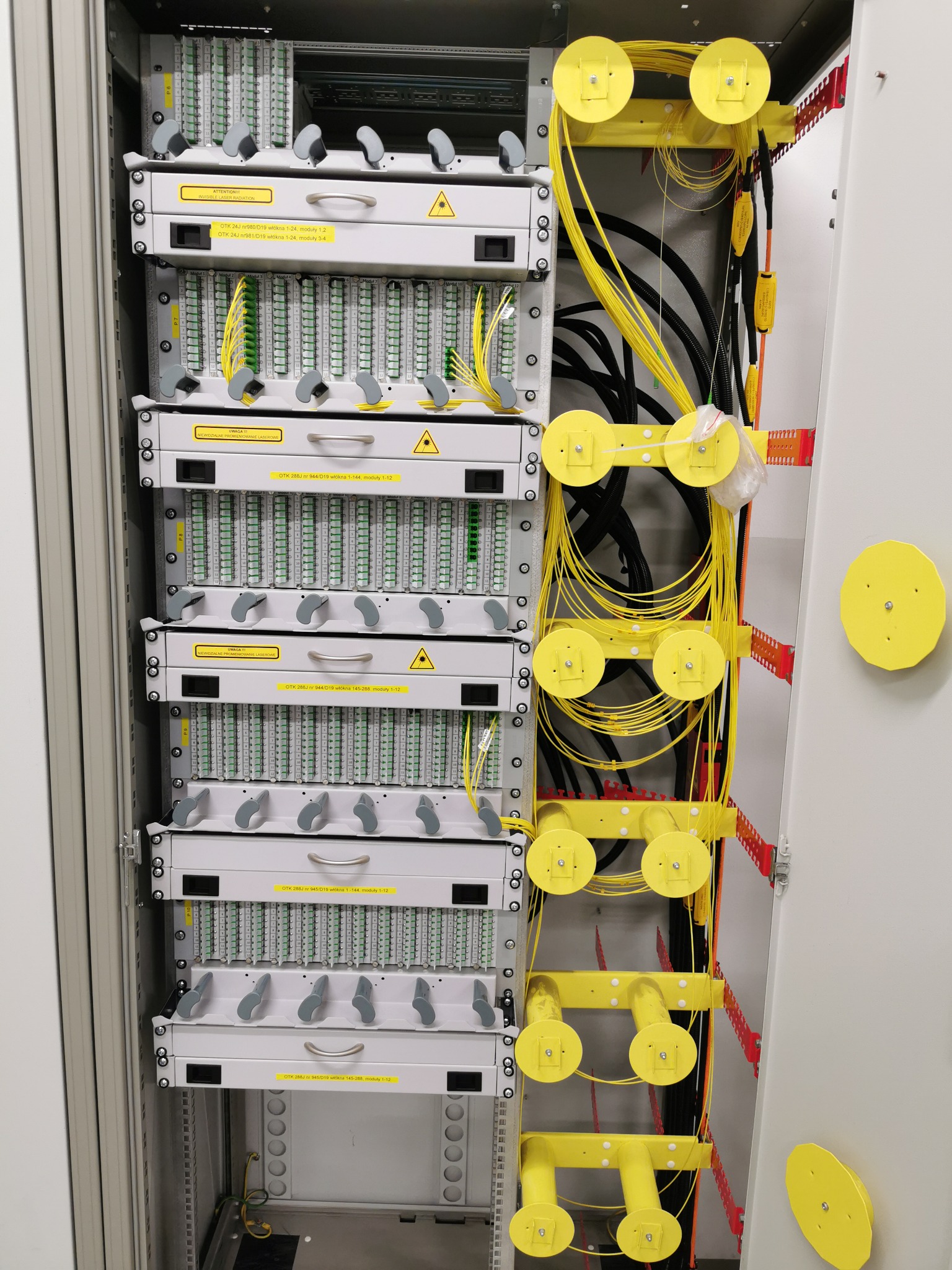 Szafa telekomunikacyjna z panelami światłowodowymi, żółtymi prowadnicami kablowymi oraz oznaczeniami ostrzegawczymi o promieniowaniu laserowym.