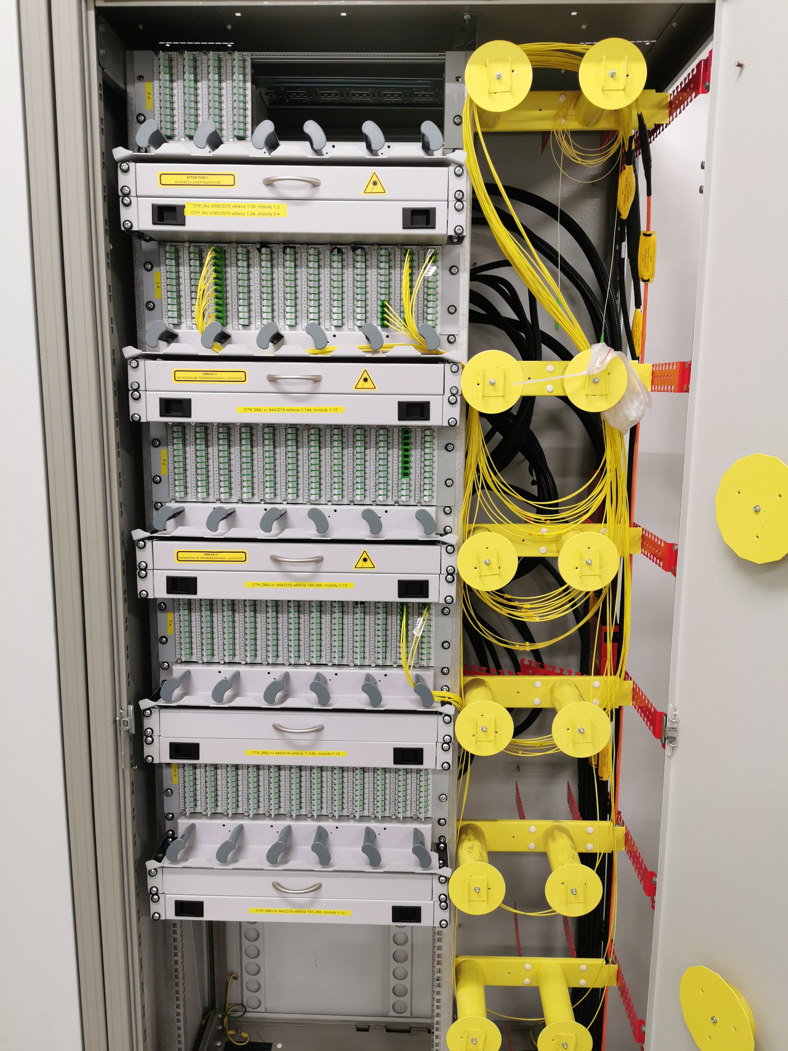 Otwarta szafa telekomunikacyjna z panelami światłowodowymi, żółtymi kablami i elementami mocującymi.
