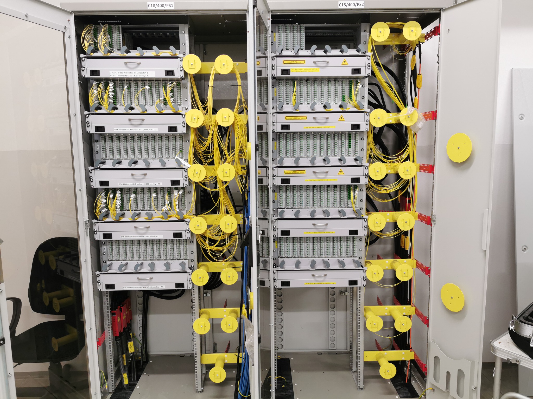 Otwarta szafa telekomunikacyjna z panelami światłowodowymi, uporządkowanymi żółtymi kablami i oznaczeniami modułów na białych etykietach.