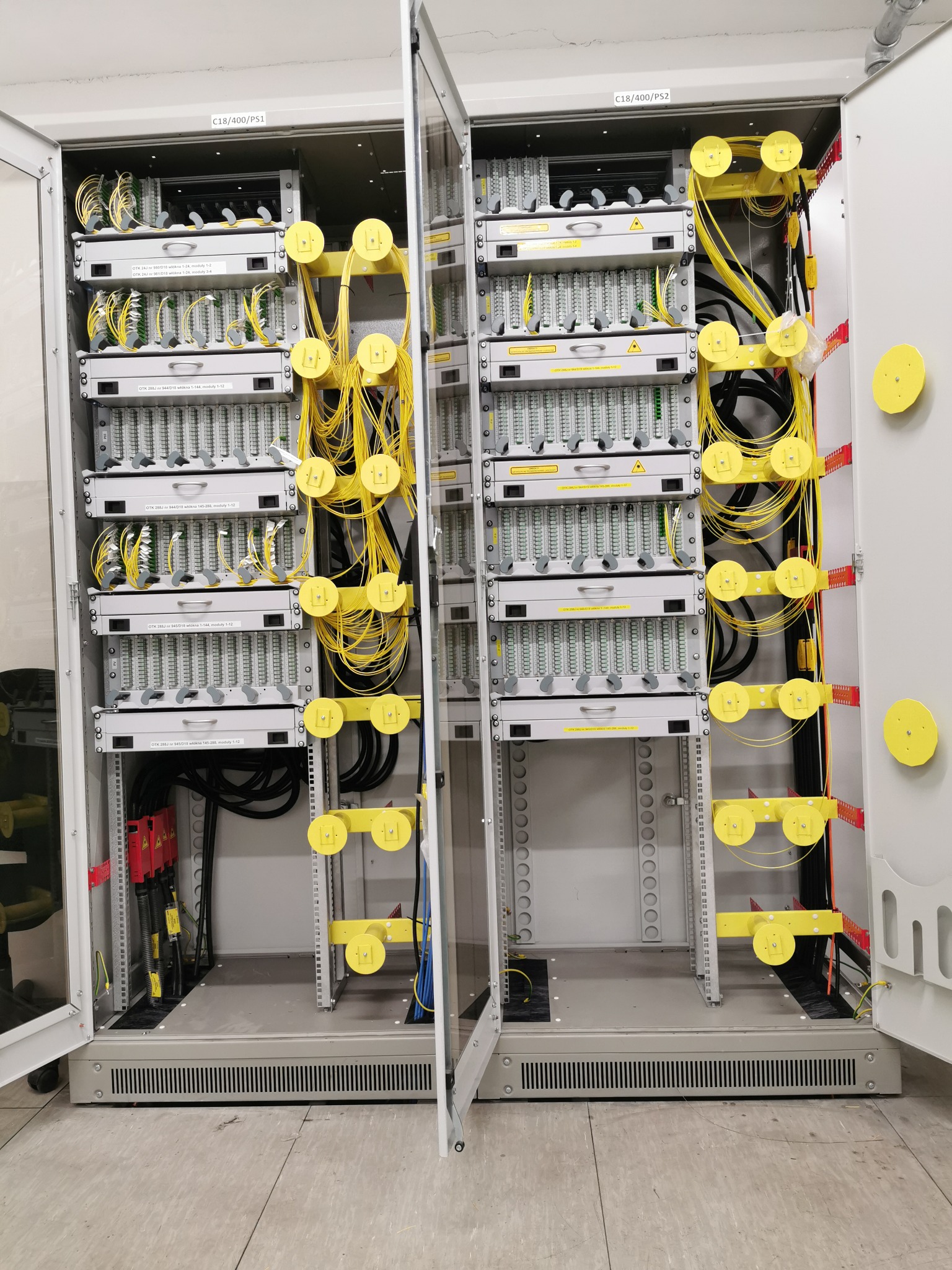 Otwarta szafa telekomunikacyjna z panelami światłowodowymi, żółtymi prowadnicami kablowymi i uporządkowanymi przewodami, widok z bliska na zainstalowany sprzęt.