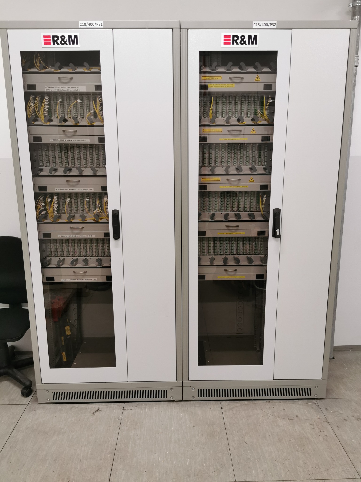 Dwie szafy rackowe z panelami krosowniczymi światłowodowymi marki R&M, widoczne kable światłowodowe i oznaczenia modułów.