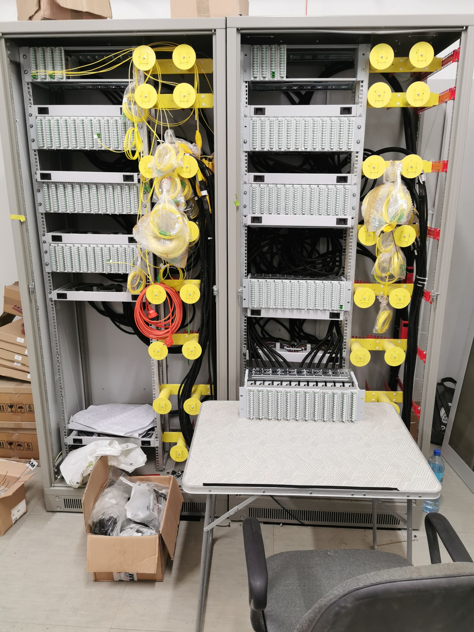 Dwa panele krosowe w szafie rack, z żółtymi elementami prowadzenia kabli, kable światłowodowe w folii, skrzynka z akcesoriami i dokumentacja na podłodze, stół roboczy z dodatkowym panelem krosowym.