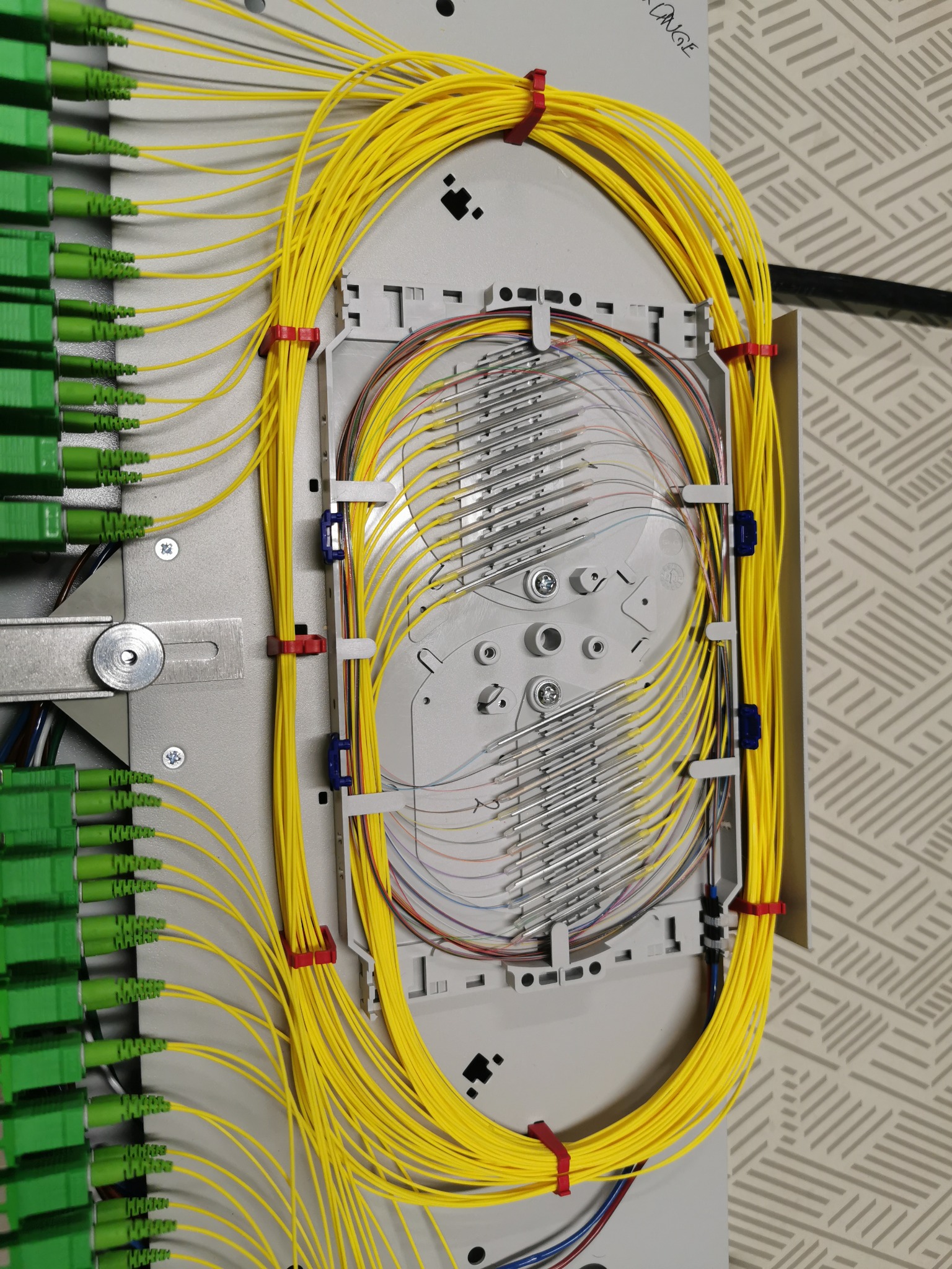 Panel światłowodowy z uporządkowanymi żółtymi kablami światłowodowymi, z zielonymi złączami po lewej stronie, zamocowanymi w szarej szafie telekomunikacyjnej, widoczny fragment ściany z wzorem w tle.