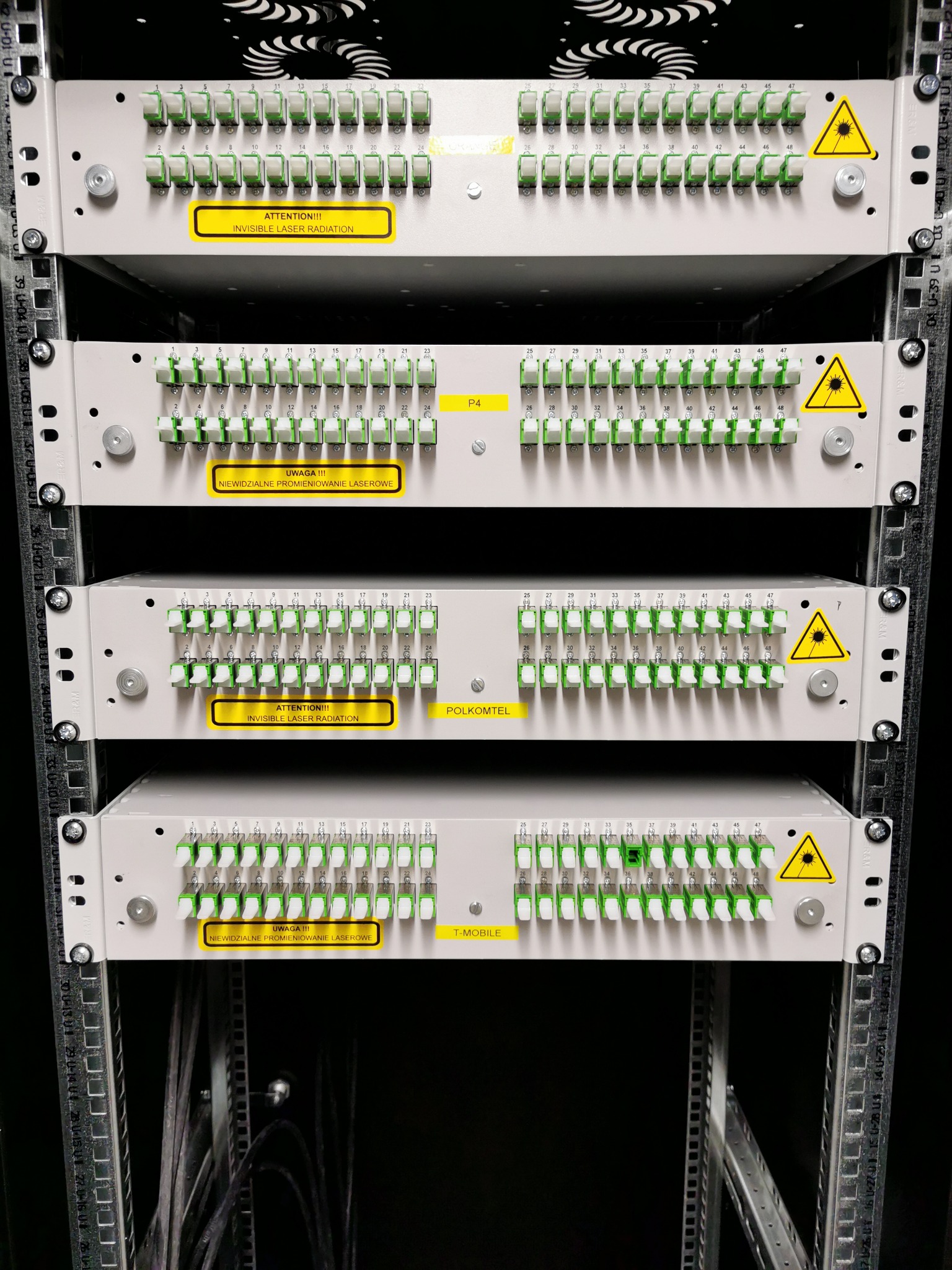 Panel światłowodowy w szafie rack z widocznymi złączami światłowodowymi, żółtymi etykietami ostrzegającymi przed promieniowaniem laserowym i oznaczeniami firm Polkomtel oraz T-Mobile.