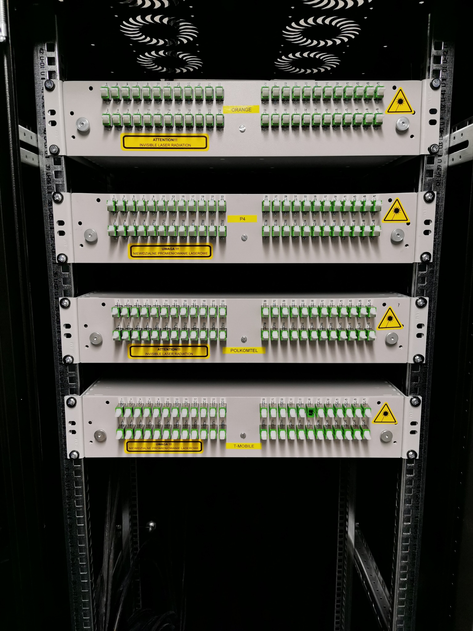 Panel krosowniczy światłowodowy w szafie rack, z widocznymi portami oznaczonymi etykietami Orange, P4, Polkomtel, T-Mobile oraz ostrzeżeniami o promieniowaniu laserowym.