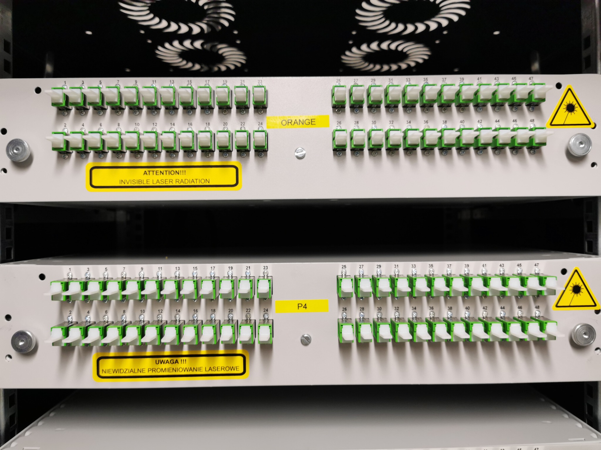 Panel krosowniczy światłowodowy z portami oznaczonymi numerami i ostrzeżeniem o promieniowaniu laserowym, zainstalowany w szafie rack.