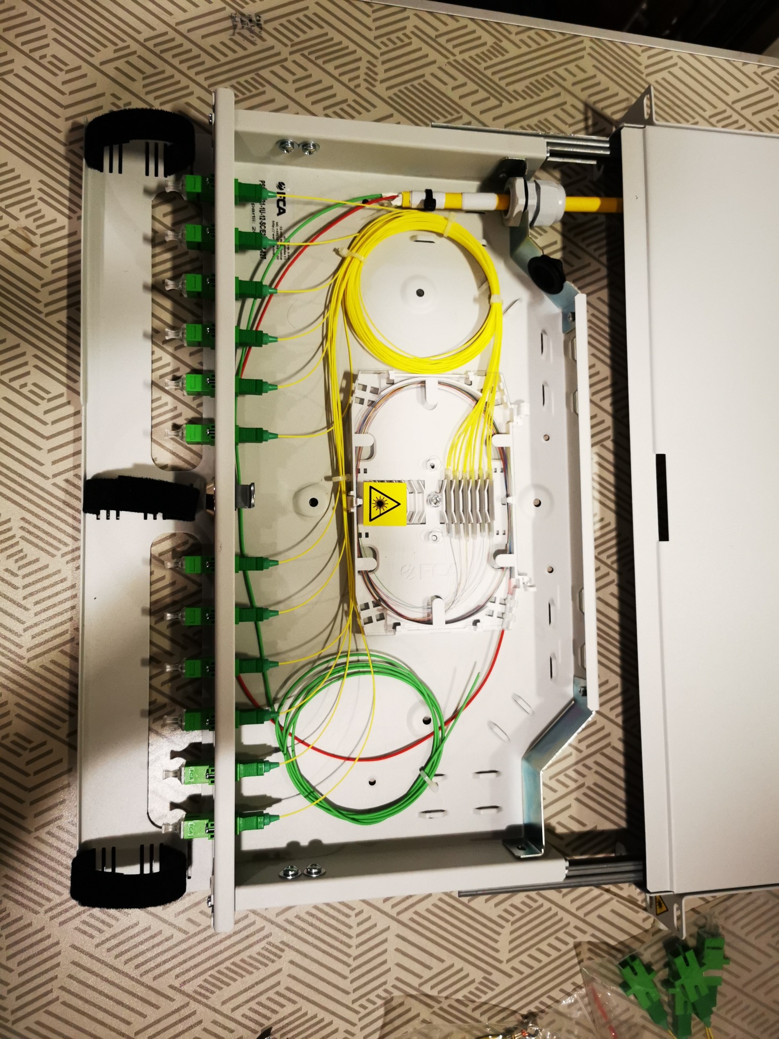 Otwarta skrzynka rozdzielcza światłowodowa z widocznymi kablami światłowodowymi w kolorach żółtym, zielonym i czerwonym, zakończonymi zielonymi złączami, z symbolem ostrzegawczym lasera.