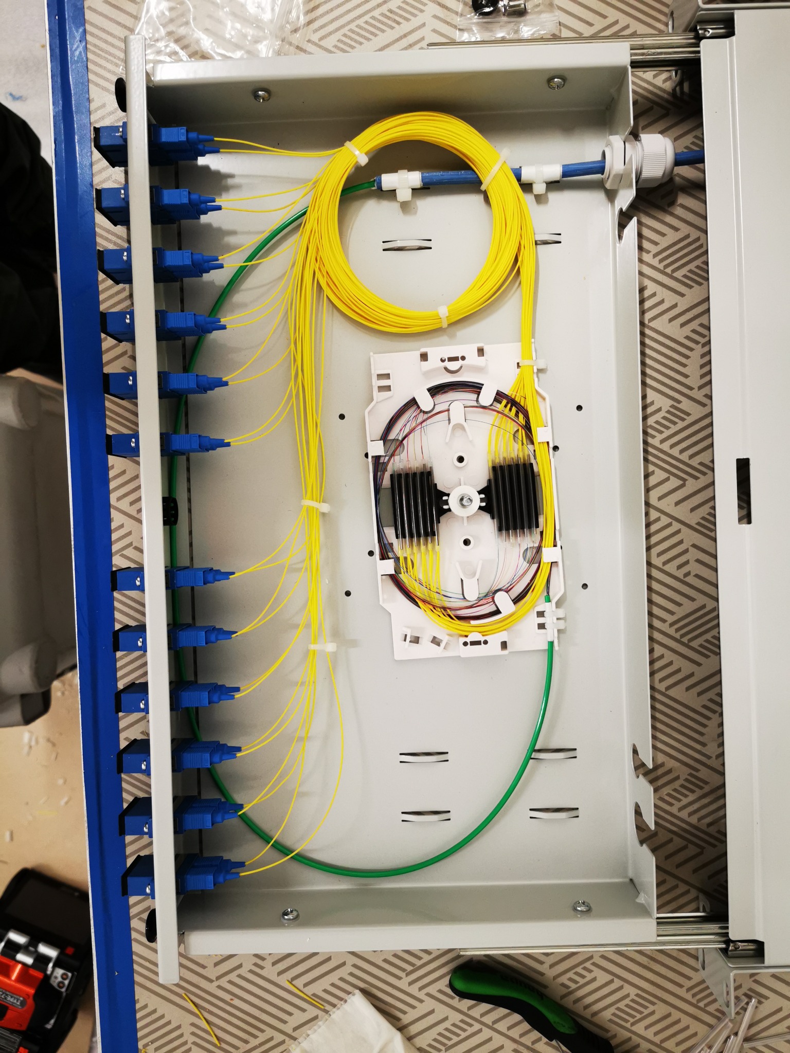 Otwarta skrzynka telekomunikacyjna z kablem światłowodowym w kolorze żółtym, podłączonym do niebieskich złączek po lewej stronie i zwiniętym wewnątrz skrzynki, widoczny element do spawania...