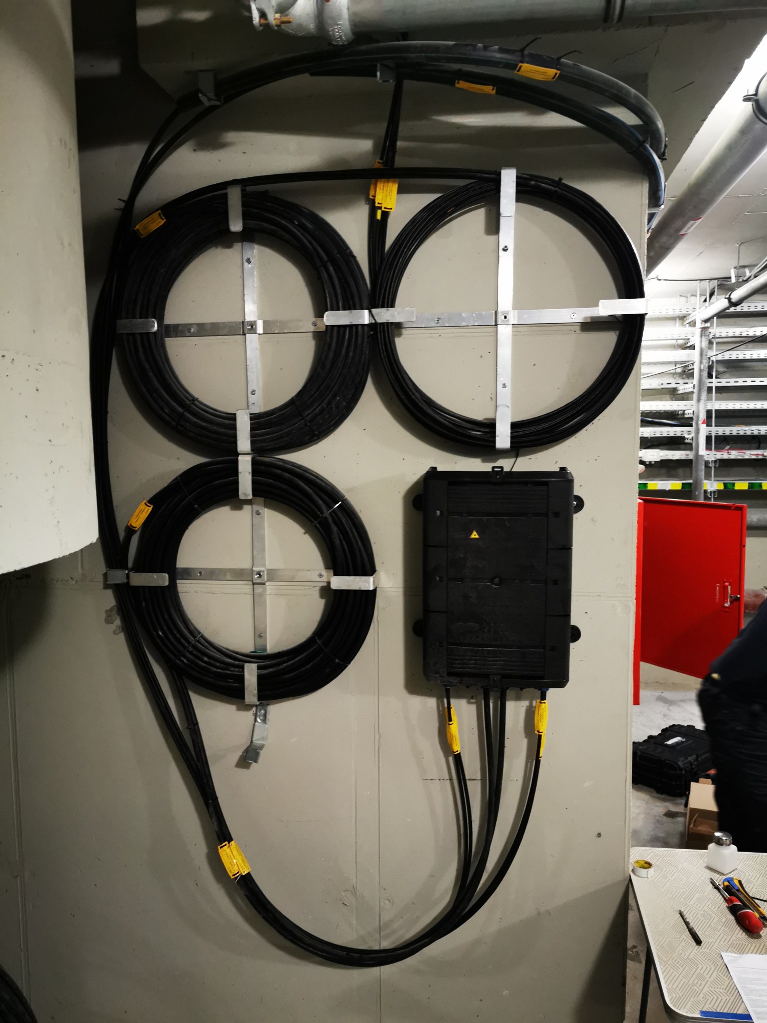 Instalacja okablowania światłowodowego: cztery zwoje kabli zamocowane na ścianie za pomocą metalowych uchwytów, obok czarna skrzynka przyłączeniowa z żółtymi oznaczeniami kabli, widoczne narzędzia...