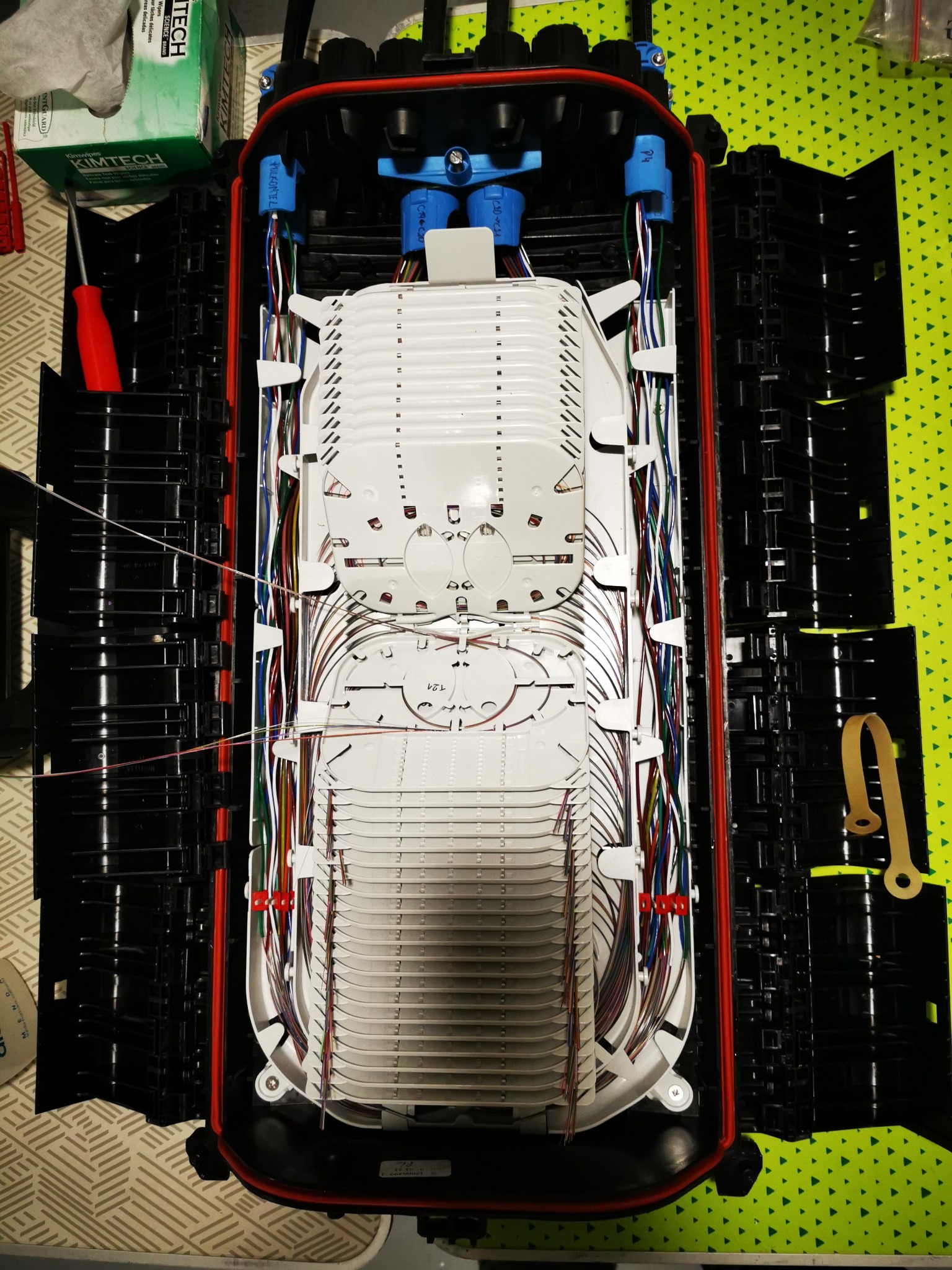 Otwarta skrzynka telekomunikacyjna z uporządkowanymi kablami światłowodowymi i elementami złącznymi, widoczna z góry, z narzędziem i pudełkiem chusteczek obok.
