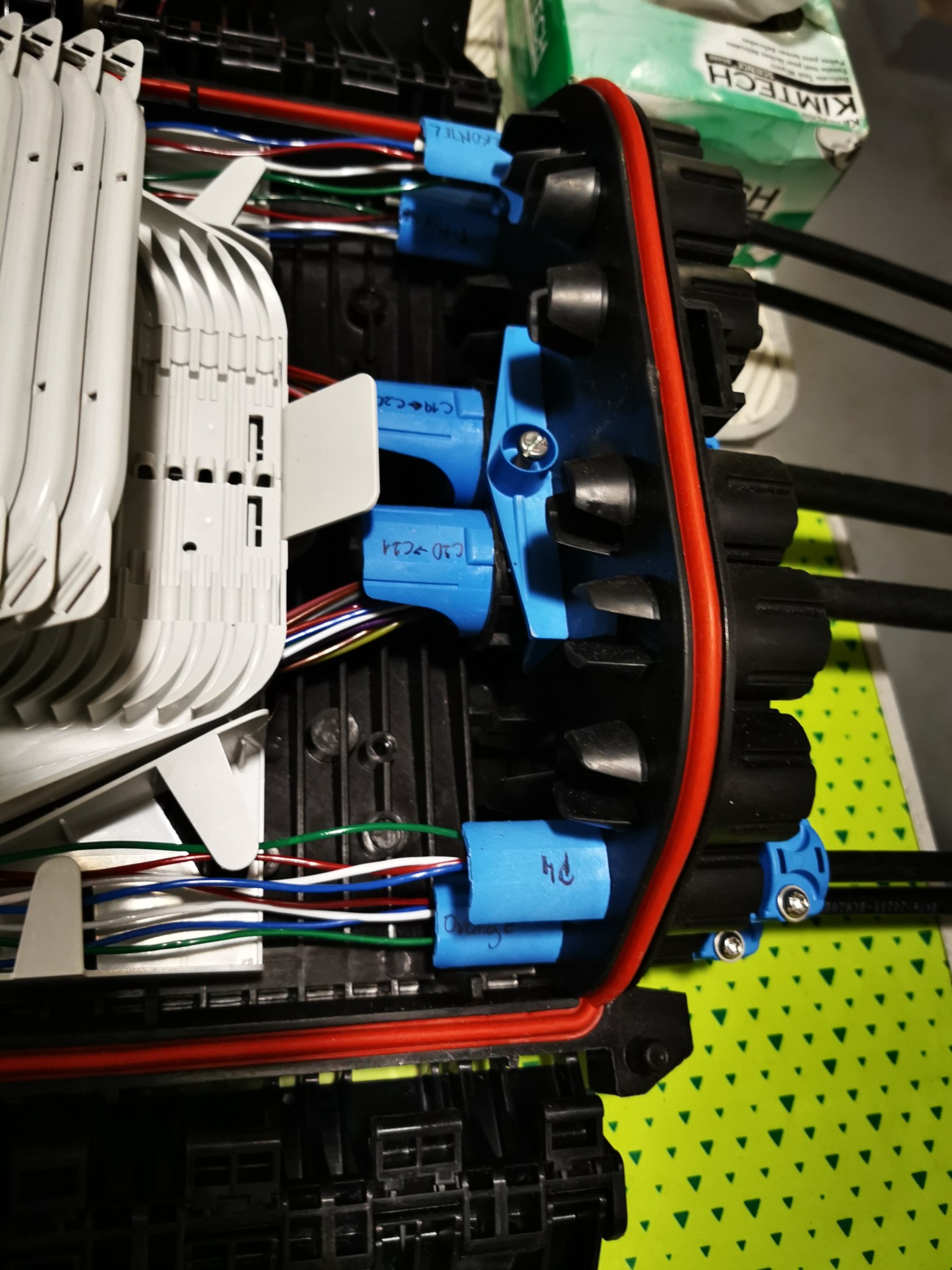 Otwarta skrzynka telekomunikacyjna z widocznymi przewodami w różnych kolorach i niebieskimi złączkami, ułożonymi wewnątrz czarnej obudowy z czerwoną uszczelką.