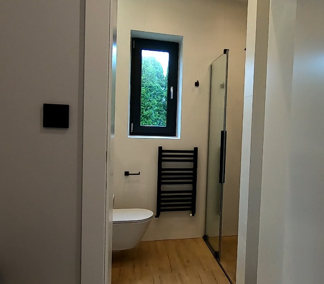 Nowoczesna łazienka z drewnianą podłogą, wiszącą toaletą, czarnym grzejnikiem drabinkowym i kabiną prysznicową ze szklanymi drzwiami, widok zza uchylonych drzwi.