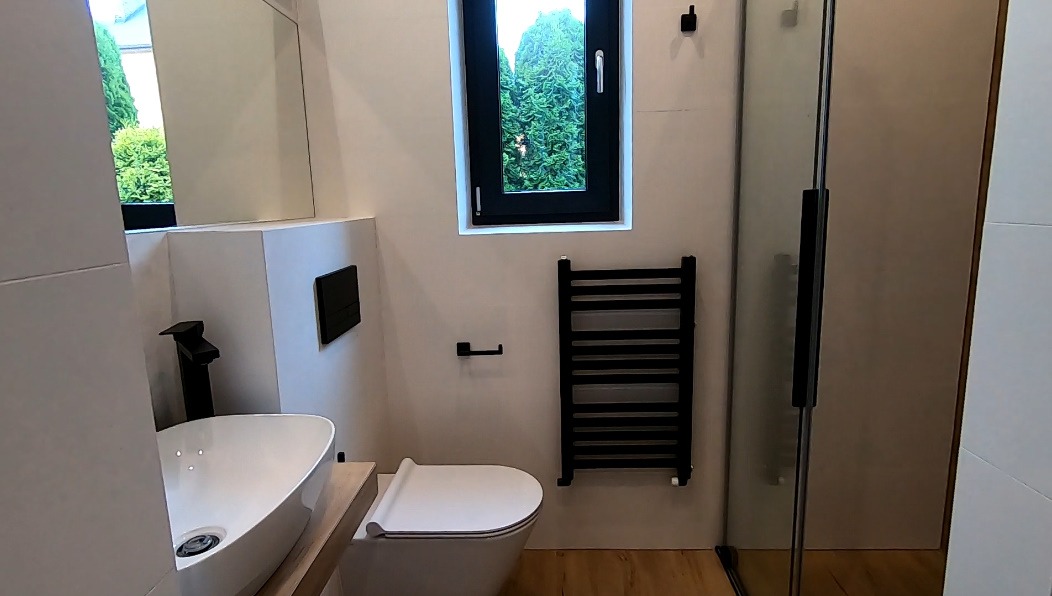 Nowoczesna łazienka z owalną umywalką nablatową, czarną armaturą, wiszącą toaletą, grzejnikiem drabinkowym i kabiną prysznicową ze szklanymi drzwiami, widok na zieleń za oknem.