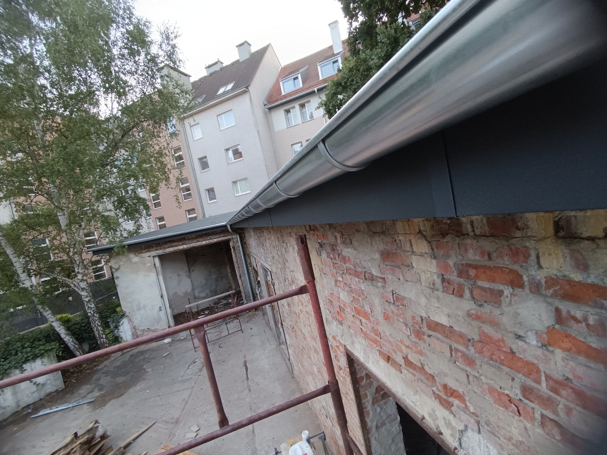 Metalowa rynna zamontowana na budynku z cegły, widok z dołu, w tle budynki mieszkalne i drzewa. Nowa rynna kontrastuje ze starą cegłą.