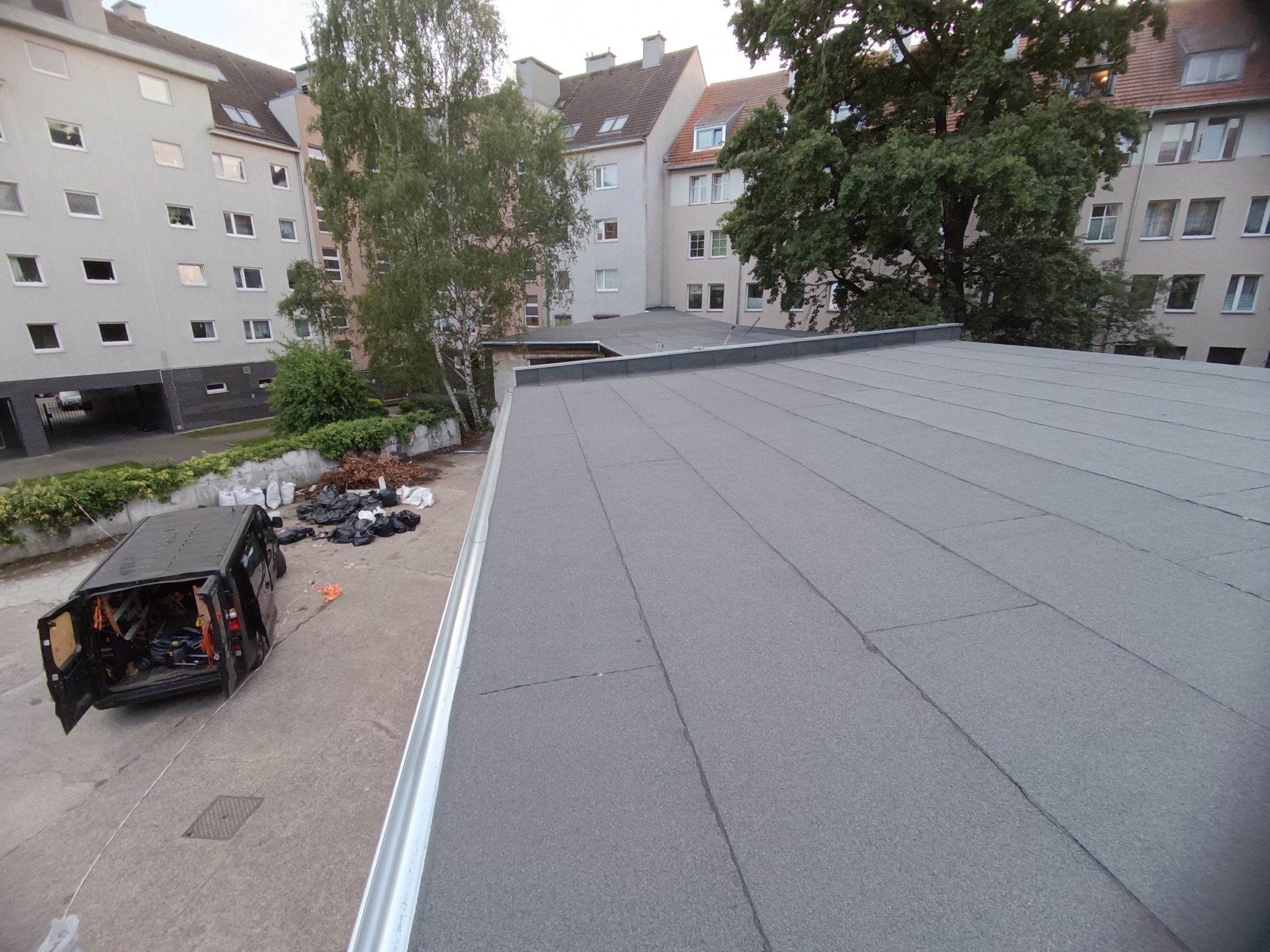Szary, płaski dach budynku wielorodzinnego po renowacji, z widoczną srebrną rynną. W tle zaparkowany bus i budynki mieszkalne.