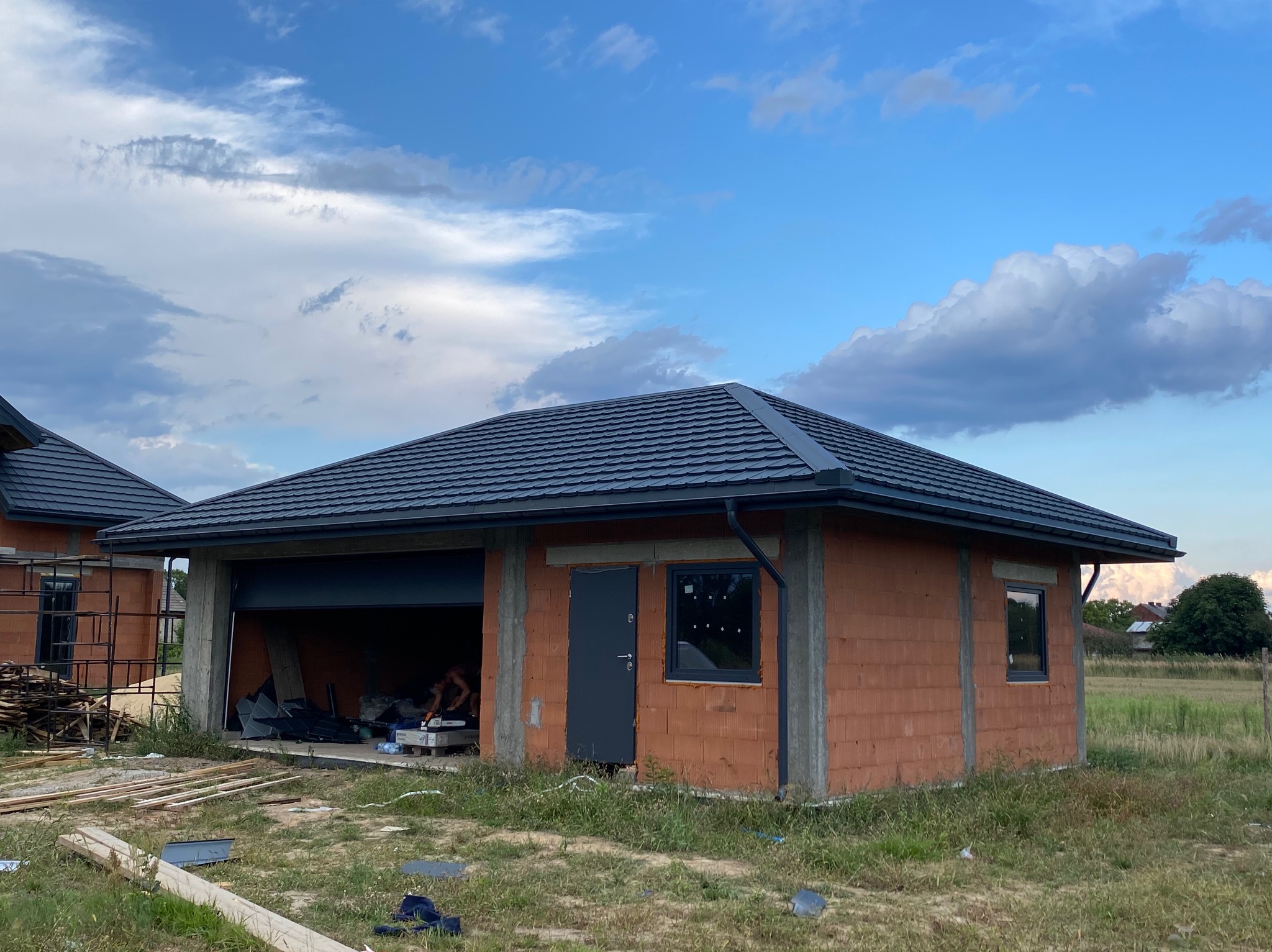 Budynek w trakcie budowy z zamontowanym, grafitowym dachem z blachy trapezowej i rynnami, widoczne cegły i betonowe słupy konstrukcyjne, otwarty garaż z materiałami budowlanymi w środku, błękitne...