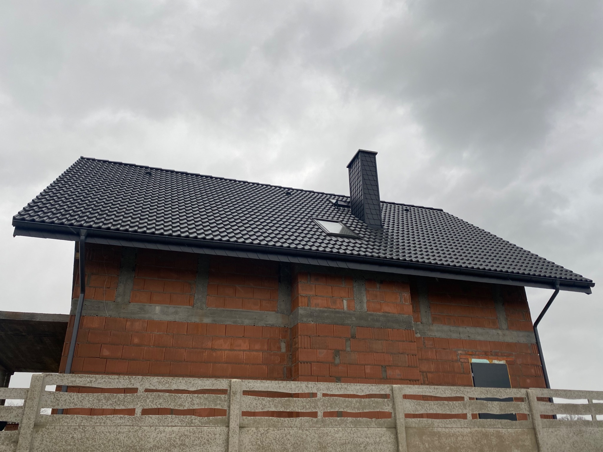 Niedokończony budynek z czarną dachówką, widok z dołu na dach z kominem i lukarną, ceglana ściana w trakcie budowy, betonowe ogrodzenie.