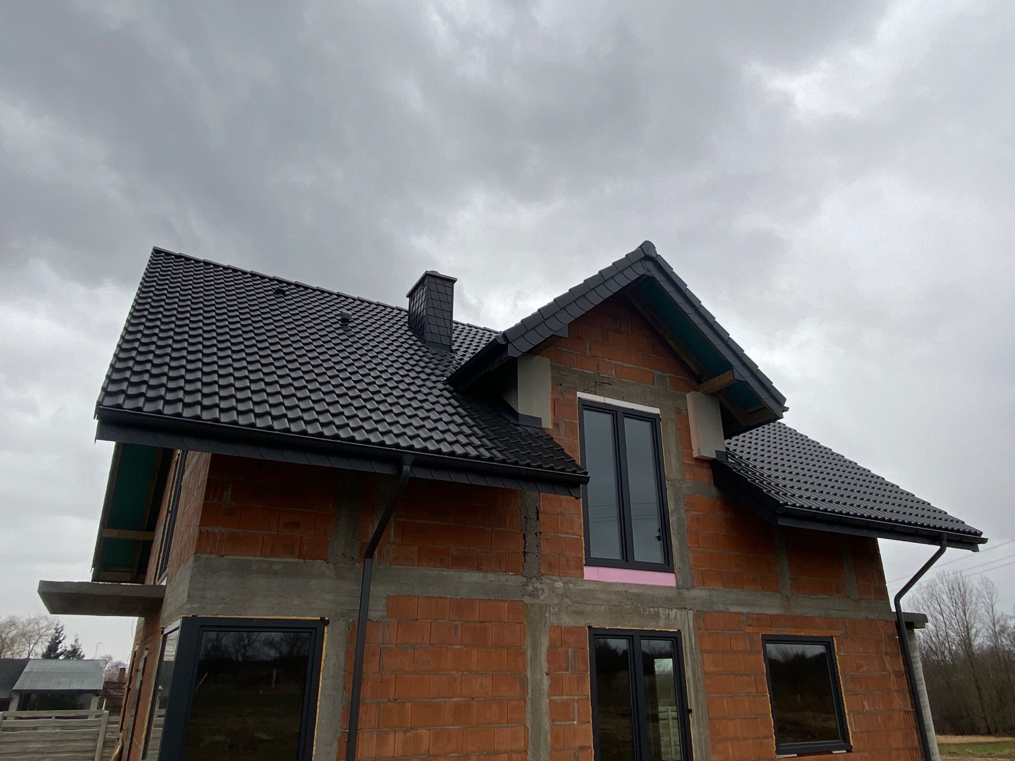 Niedokończony budynek z cegły z zamontowanym czarnym dachem z blachodachówki i czarnymi rynnami, widoczny komin z ciemnej dachówki, w tle pochmurne niebo.
