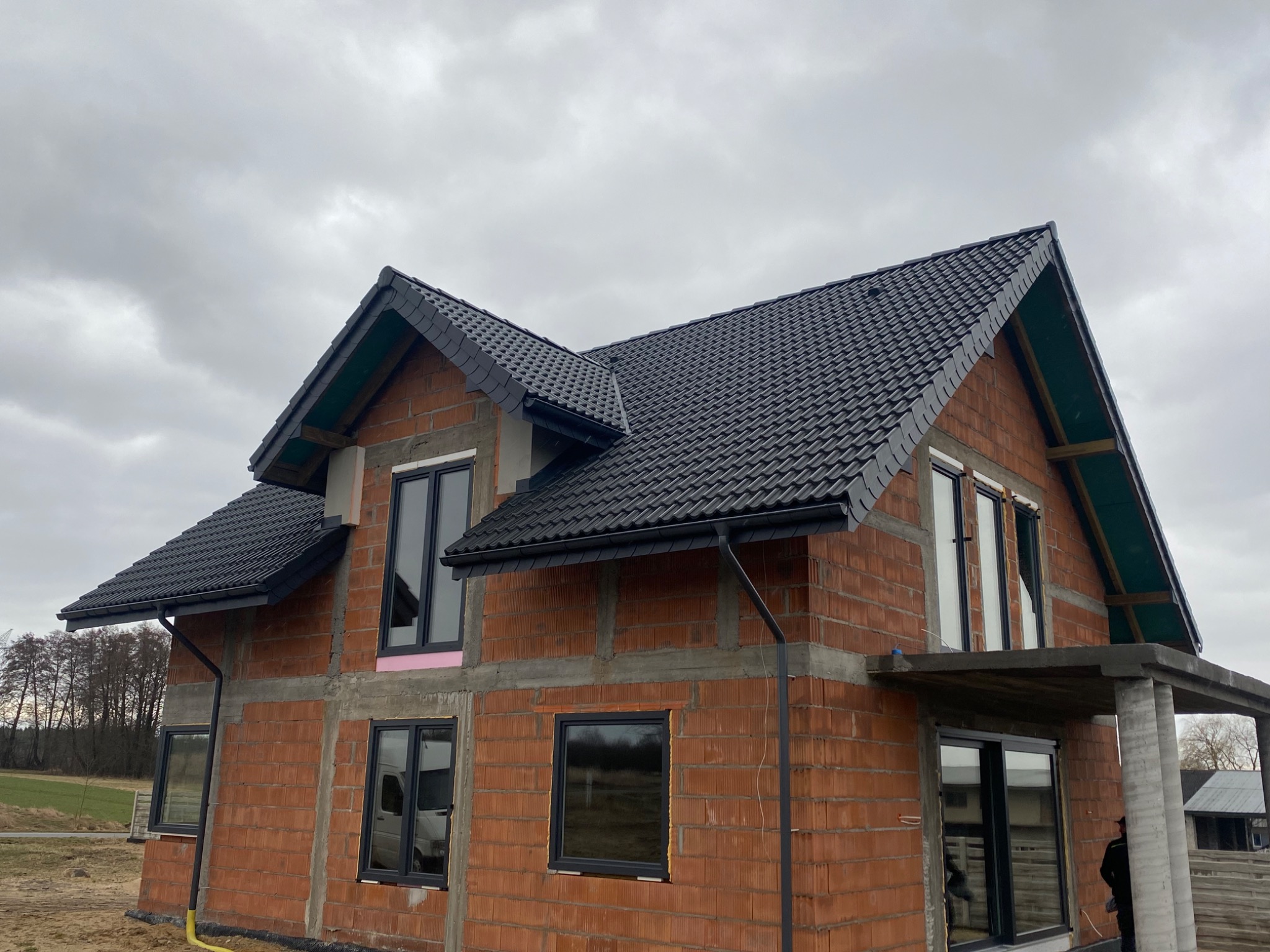 Dom w trakcie budowy z zamontowaną czarną dachówką i orynnowaniem, widoczna surowa cegła i betonowe elementy konstrukcyjne, różowy styropian izolacyjny przy oknie.