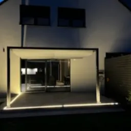 Nowoczesna pergola z białym zadaszeniem i oświetleniem LED wzdłuż krawędzi tarasu, na tle białej ściany domu, nocne ujęcie.