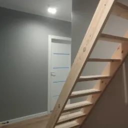 Surowe drewniane schody strychowe zamontowane w szarej ścianie, prowadzące do otworu w suficie wykończonym płytą karton gips, widoczne rury instalacyjne pod sufitem.