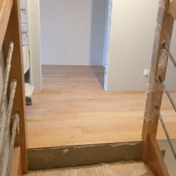 Tob-Rem Tobiasz Żmud - Widok na drewniane schody z metalowymi prętami zabezpieczonymi folią, prowadzące do pomieszczenia z jasną podłogą i szarą ścianą z gniazdkiem elektrycznym, w trakcie remontu.