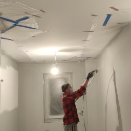 Tob-Rem Tobiasz Żmud - Mężczyzna w czapce i masce maluje ściany w pomieszczeniu na biało za pomocą pistoletu natryskowego, okno i lampa zabezpieczone folią, widoczne taśmy malarskie na suficie.