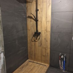 Tob-Rem Tobiasz Żmud - Kabina prysznicowa z wykończeniem imitującym drewno na ścianie i podłodze, czarna armatura, szare płytki na pozostałych ścianach i niezabudowane rury doprowadzające wodę.