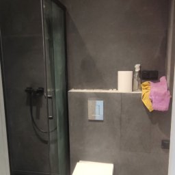 Tob-Rem Tobiasz Żmud - Nowoczesna łazienka w odcieniach szarości z kabiną prysznicową typu walk-in, podwieszaną toaletą z chromowanym przyciskiem spłukiwania i akcesoriami łazienkowymi na półce.
