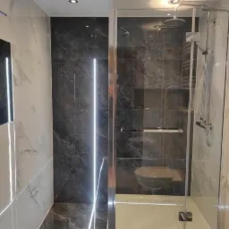 Nowoczesna łazienka z kabiną prysznicową typu walk-in, wykończona płytkami imitującymi marmur w odcieniach szarości i bieli, z akcentem w postaci pionowego oświetlenia LED na ścianie.