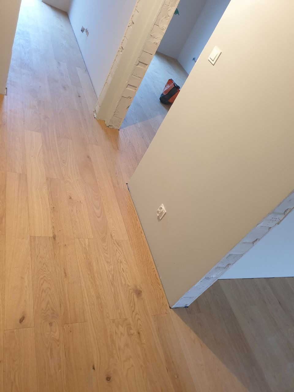 Widok na świeżo położoną drewnopodobną podłogę w korytarzu, z widocznym fragmentem ściany z gniazdkiem i niedokończonym ościeżem drzwiowym.