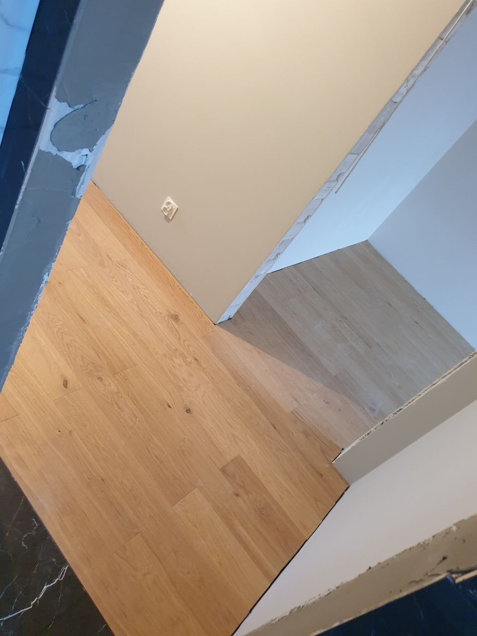 Fragment remontowanego pomieszczenia z drewnianą podłogą, świeżo pomalowaną ścianą w odcieniach bieli i gniazdkiem elektrycznym, widoczne ślady prac wykończeniowych przy listwach przypodłogowych.