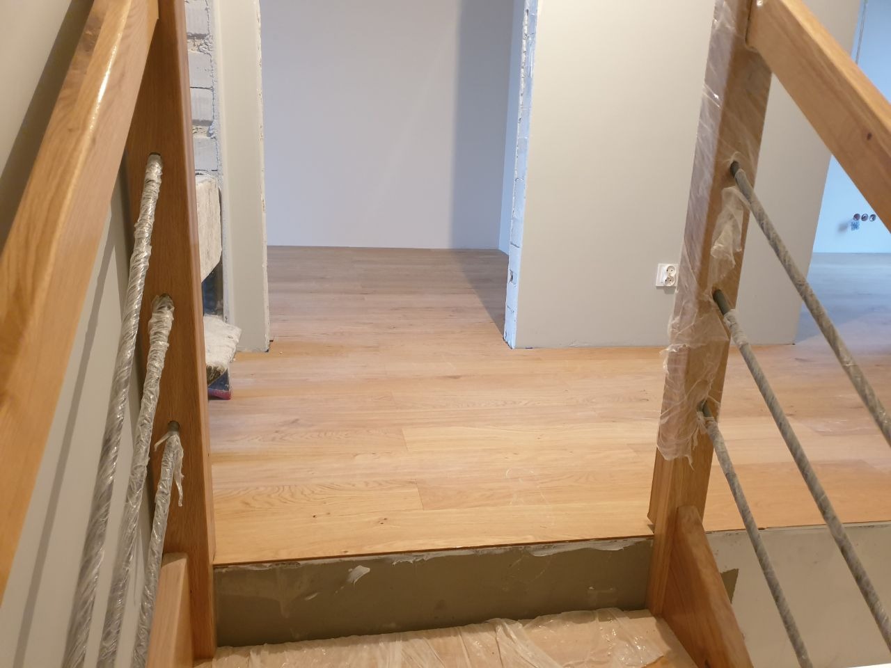 Widok na drewniane schody z metalowymi prętami zabezpieczonymi folią, prowadzące do pomieszczenia z jasną podłogą i szarą ścianą z gniazdkiem elektrycznym, w trakcie remontu.