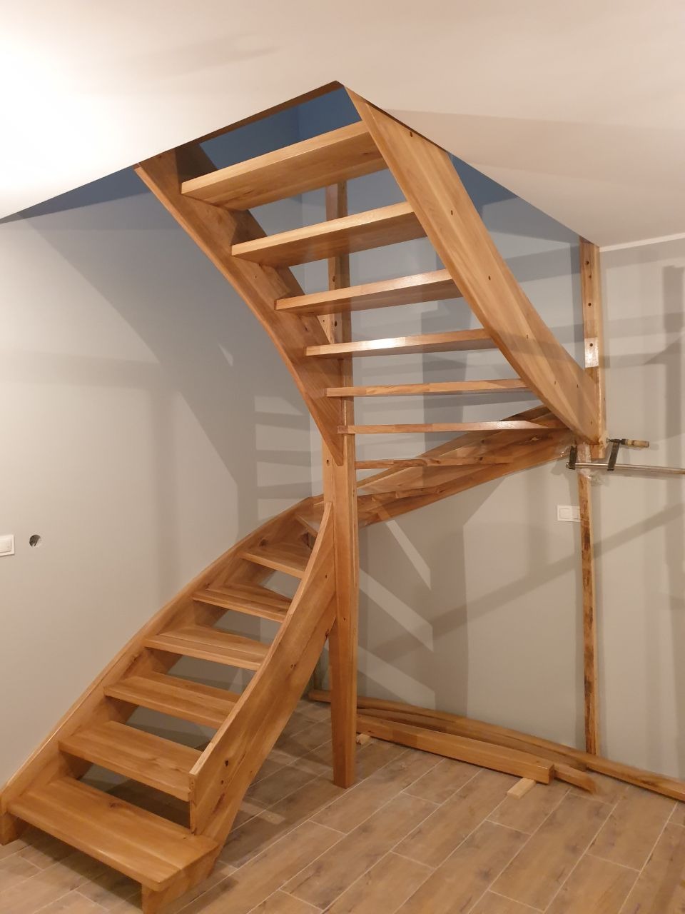 Drewniane schody z jasnego drewna, częściowo zamontowane, widoczne deski podłogowe i fragment ściany w szarym odcieniu.