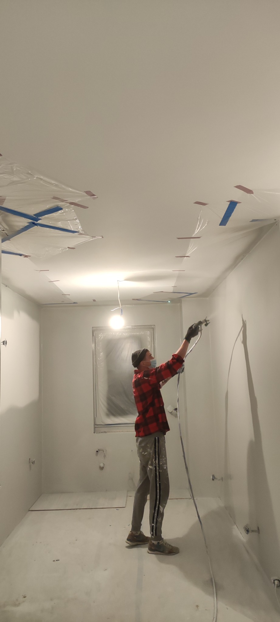 Mężczyzna w czapce i masce maluje ściany w pomieszczeniu na biało za pomocą pistoletu natryskowego, okno i lampa zabezpieczone folią, widoczne taśmy malarskie na suficie.