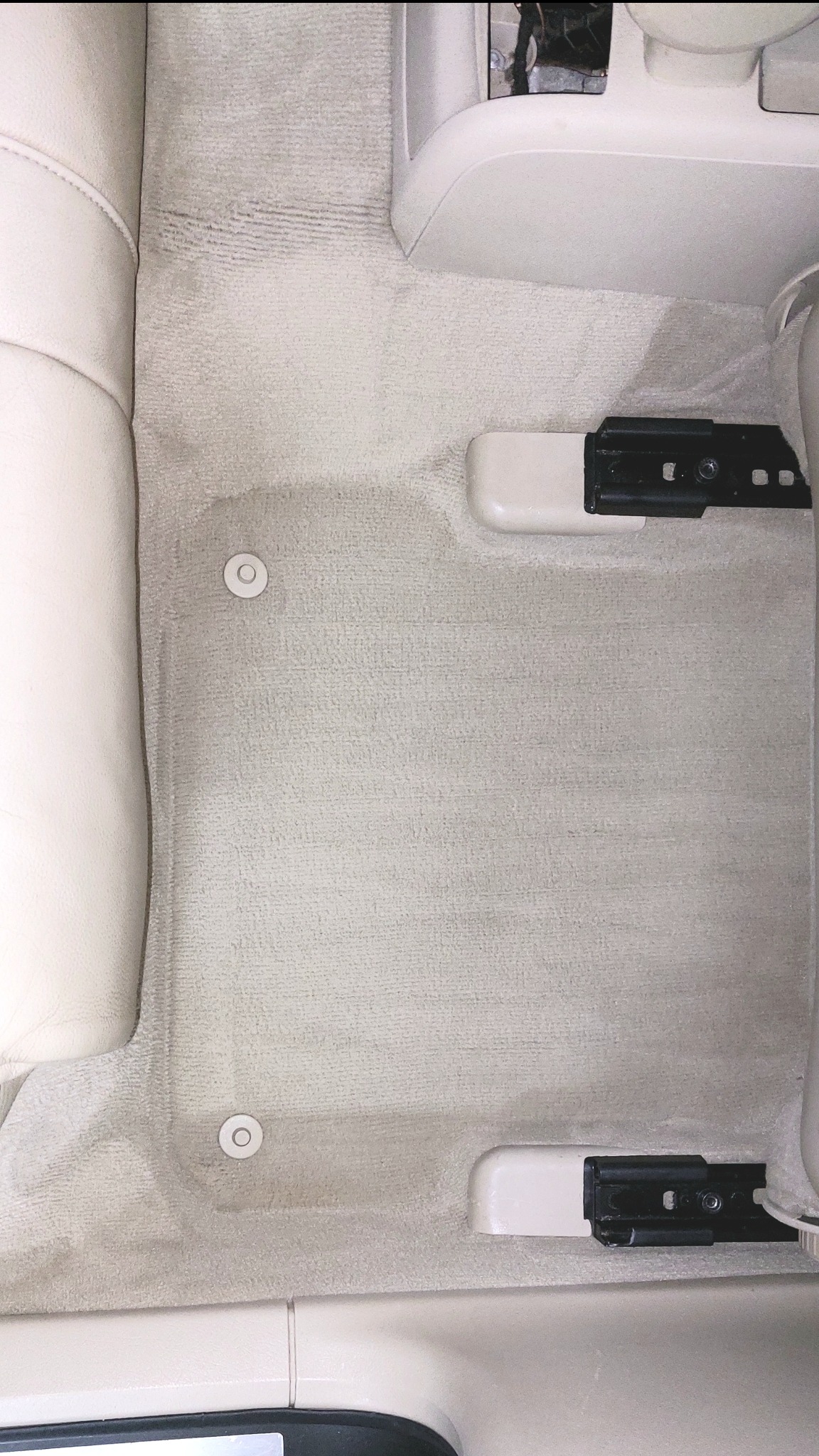 Jasna, beżowa wykładzina podłogowa w samochodzie po czyszczeniu, widoczne mocowania foteli i obudowa, ślady lekkich zabrudzeń.