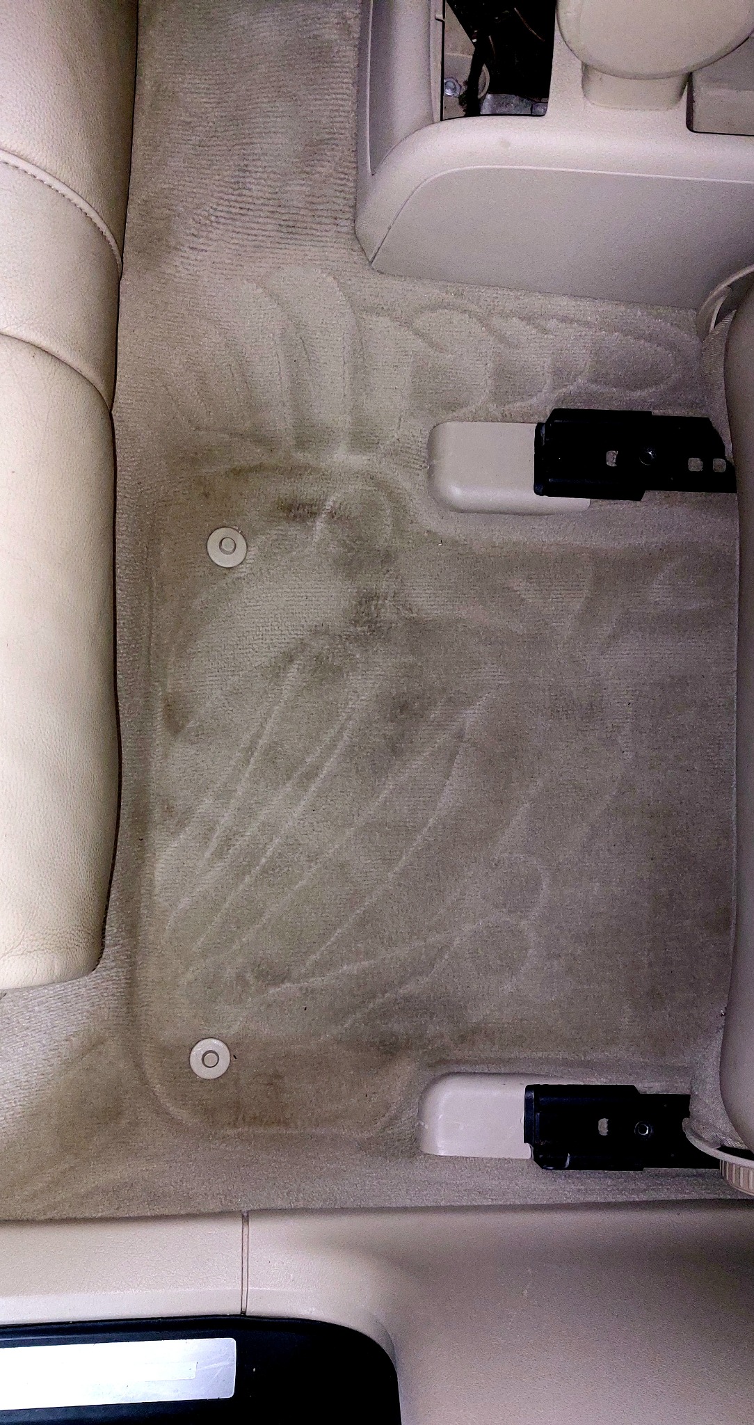 Beżowa wykładzina podłogowa samochodu z widocznymi śladami użytkowania i plamami przed czyszczeniem, widoczne elementy mocujące dywaniki.