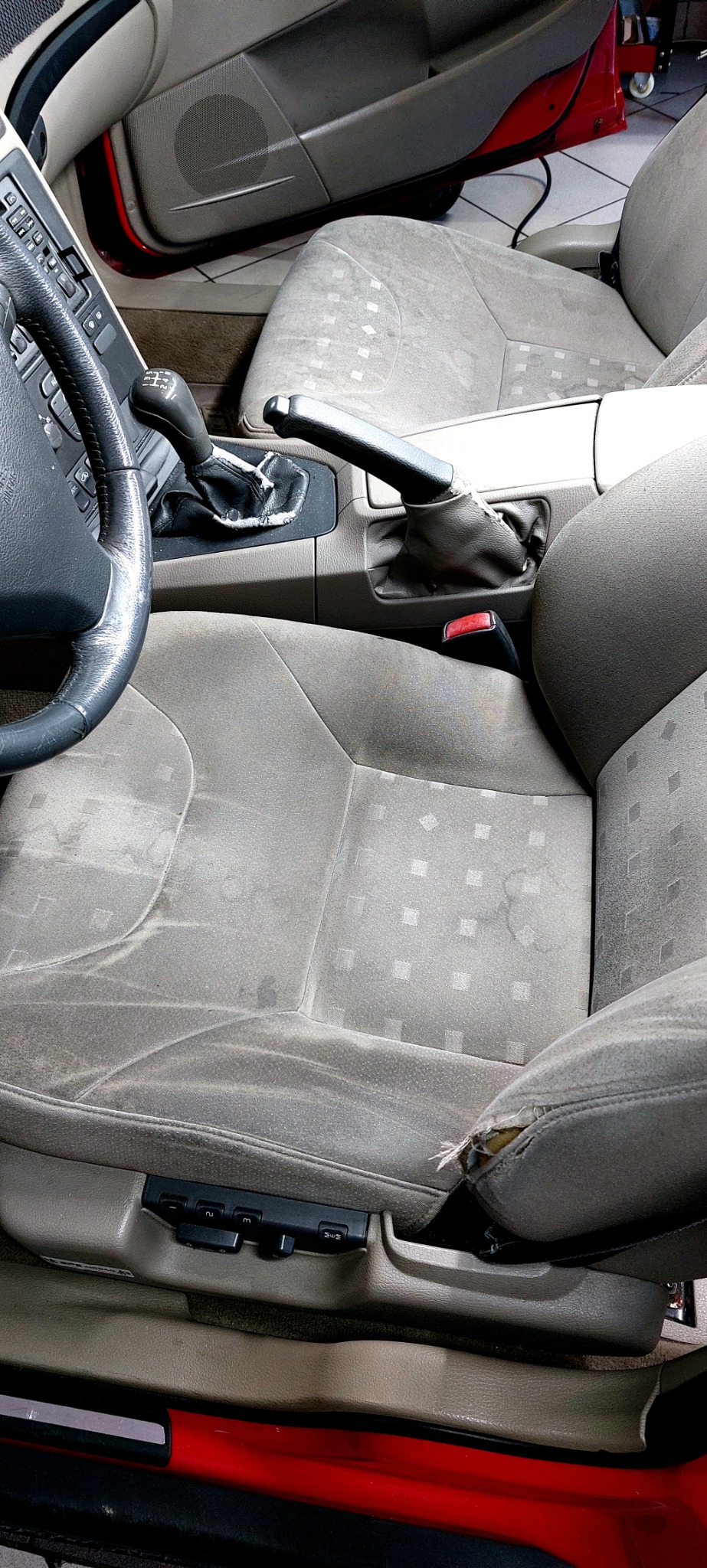 Bardzo brudne, szare siedzenie samochodowe z dziurą w boku i plamami na tapicerce, widoczna kierownica, dźwignia zmiany biegów i hamulec ręczny.