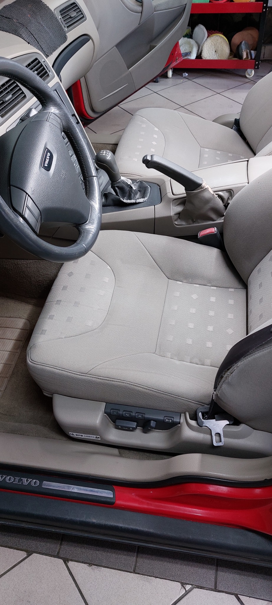 Wnętrze samochodu Volvo z beżową tapicerką, widoczne zabrudzenia na fotelu kierowcy i uszkodzona skóra wokół dźwigni zmiany biegów.