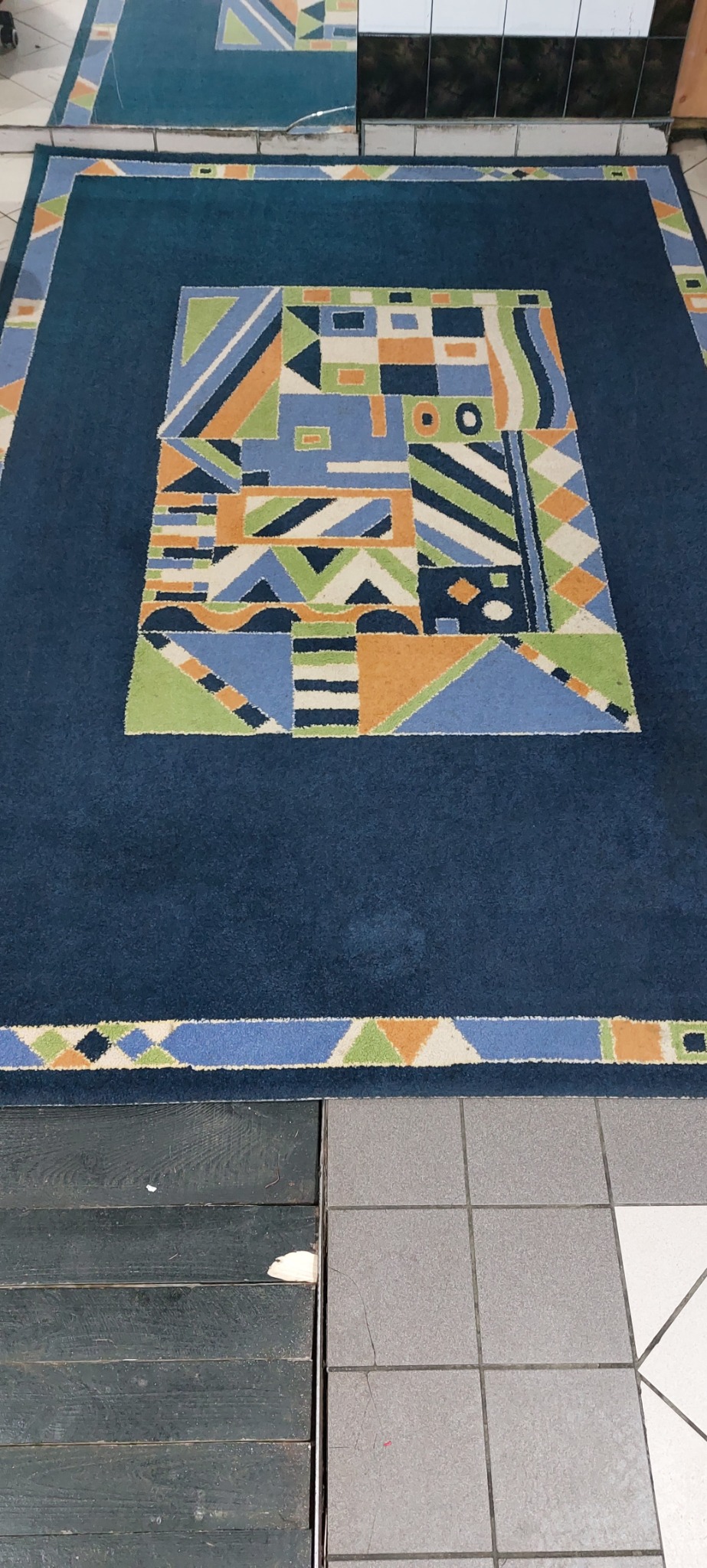 Ciemnoniebieski dywan z geometrycznym wzorem w odcieniach błękitu, zieleni i pomarańczy, leżący na styku drewnianej podłogi i szarej płytki podłogowej.