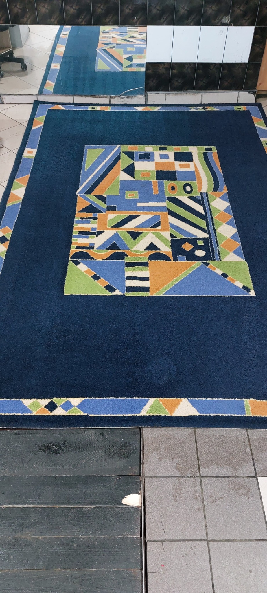 Granatowy dywan z geometrycznym wzorem w odcieniach zieleni, niebieskiego i pomarańczowego, leżący na podłodze z płytek.