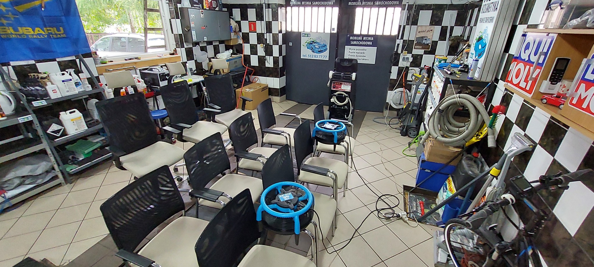 Wnętrze myjni samochodowej z krzesłami przygotowanymi do suszenia tapicerki za pomocą wentylatorów DRI POD, w tle widoczne regały z detergentami i sprzęt do czyszczenia.
