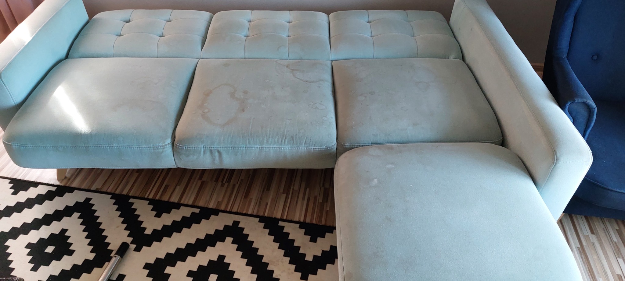 Jasna, narożna kanapa z widocznymi plamami na siedziskach, stojąca na drewnianej podłodze obok czarno-białego dywanu w geometryczne wzory.