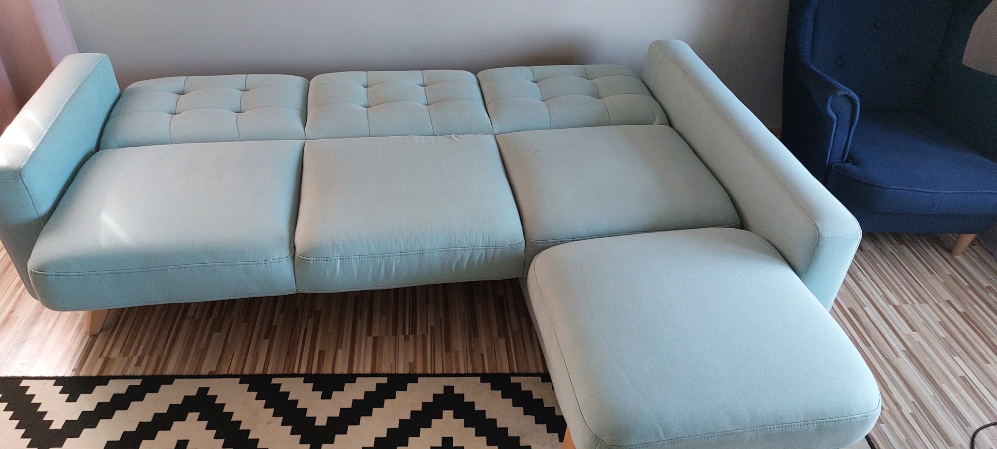 Rozłożona, jasnoniebieska sofa narożna z pikowanymi poduchami, na drewnianej podłodze z dywanem w zygzaki, obok granatowy fotel.