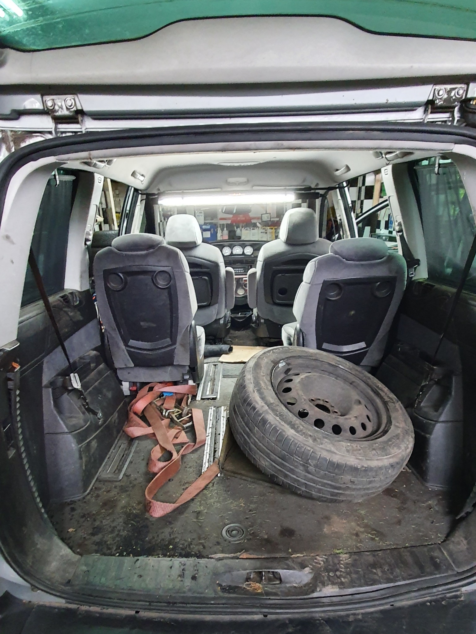 Wnętrze bagażnika samochodu osobowego z widocznymi szarymi siedzeniami, brudną podłogą, oponą i pasami transportowymi w warsztacie.